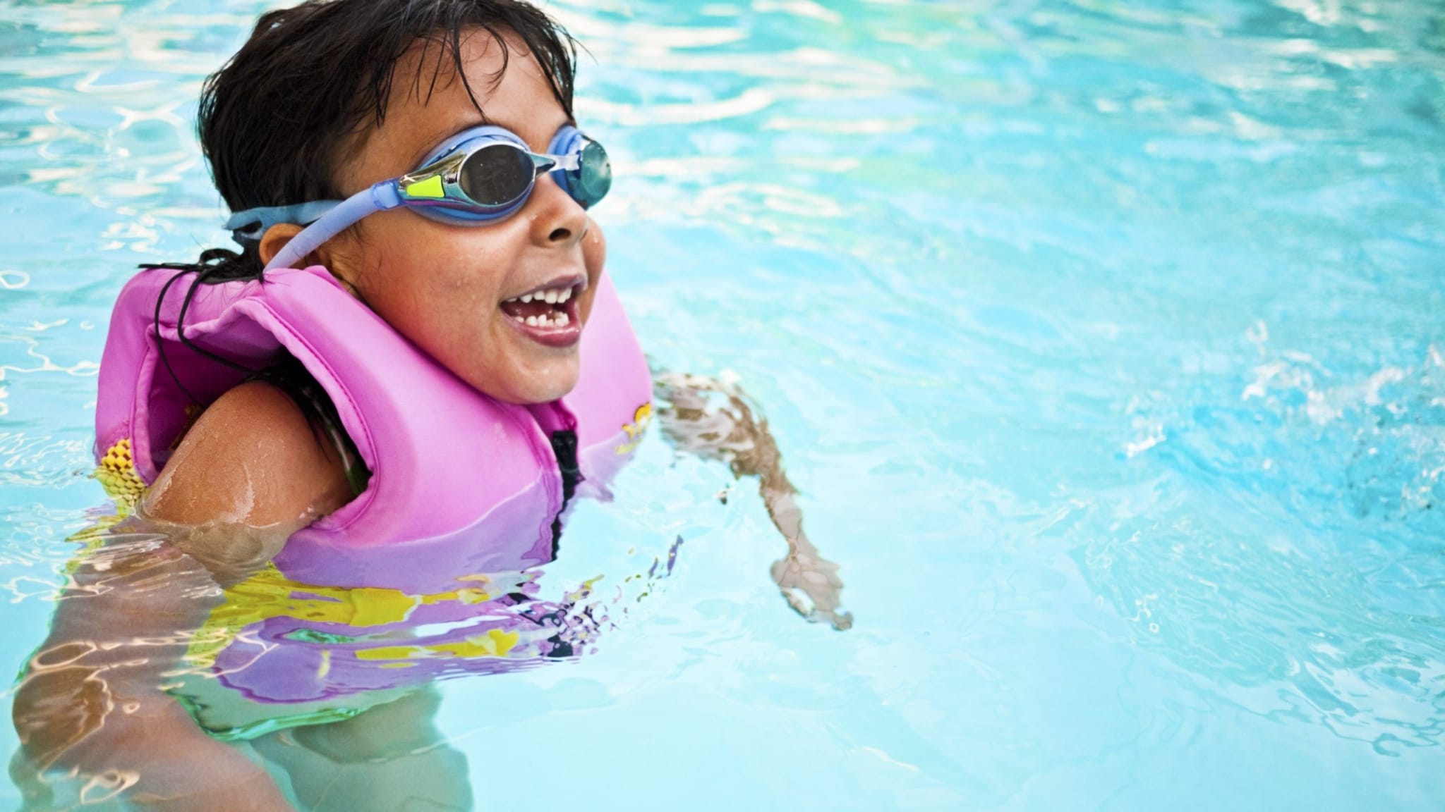 Young boy in pool wearing a lifejacket and goggles.