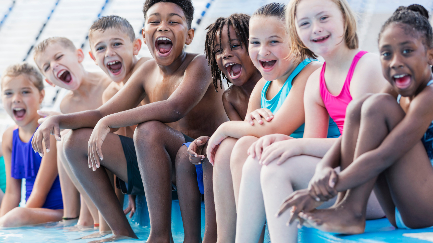 Kids at edge of pool resized A group of ethnically diverse children sitting in a line at the edge of a pool in their bathing suits laughing.